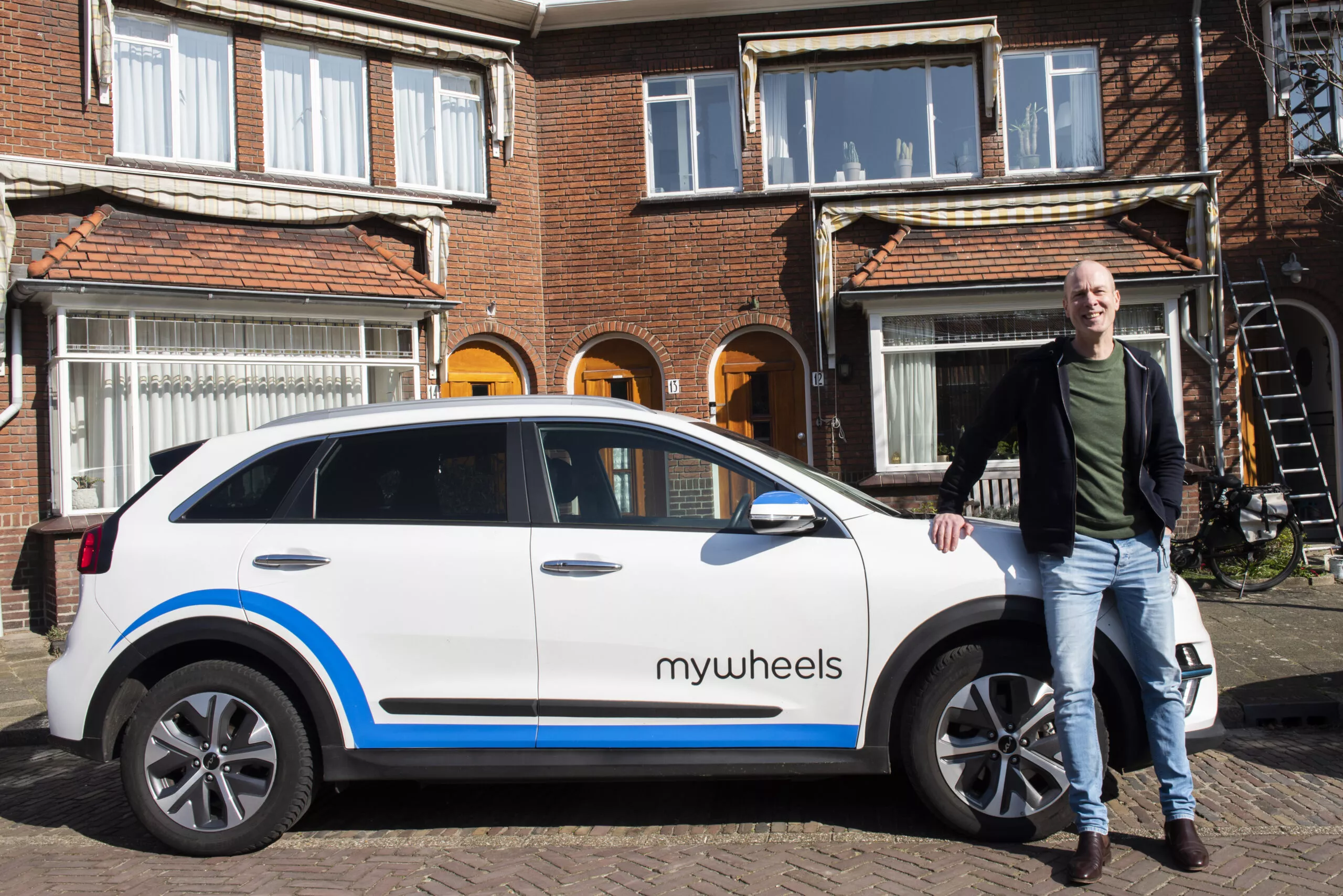 Foto van Sander waar hij glimlachend voor een deelauto staat genaamd Mywheels.