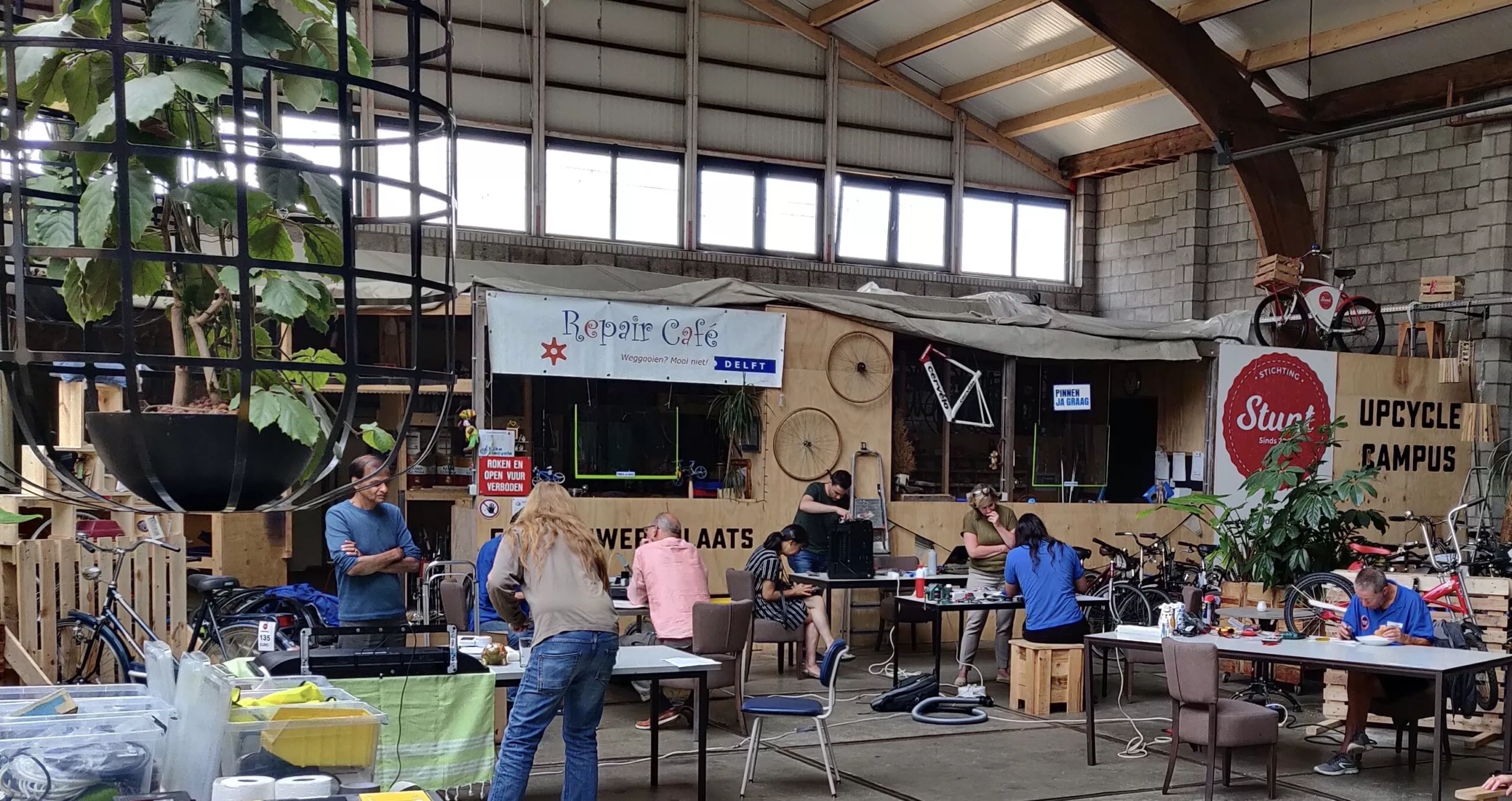 De werkplaats van het repair cafe is te zien met meerdere volwassenen in beeld, iedereen werkt in groepjes aan een project.