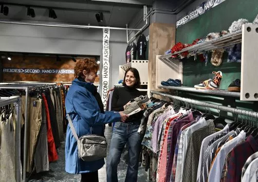 vrouw met blauwe jas en vrouw met zwarte trui staan bij een kledingrek in de winkel.