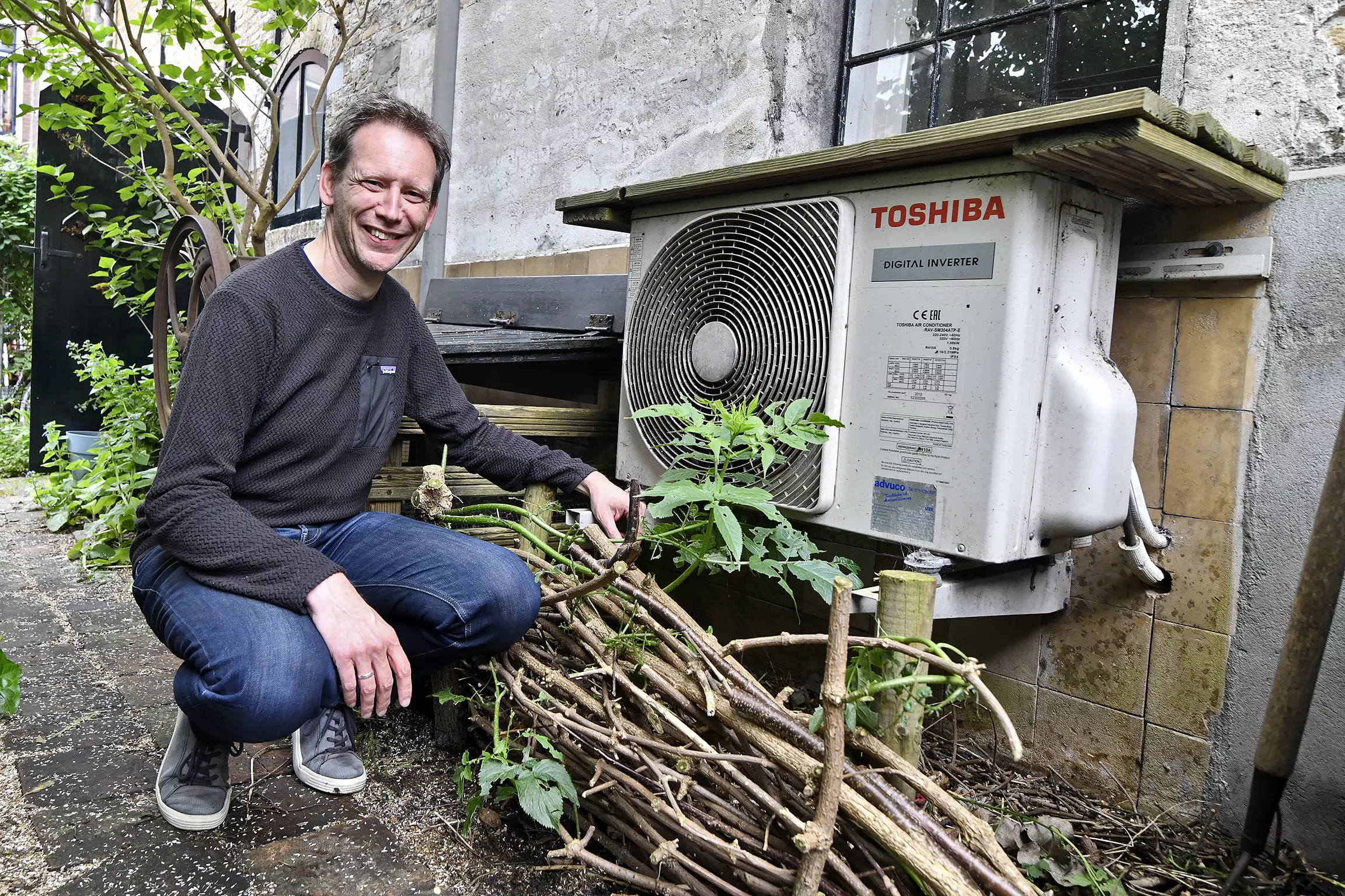 man met zwarte trui zit gehurkt bij een buitenunit van een warmtepomp