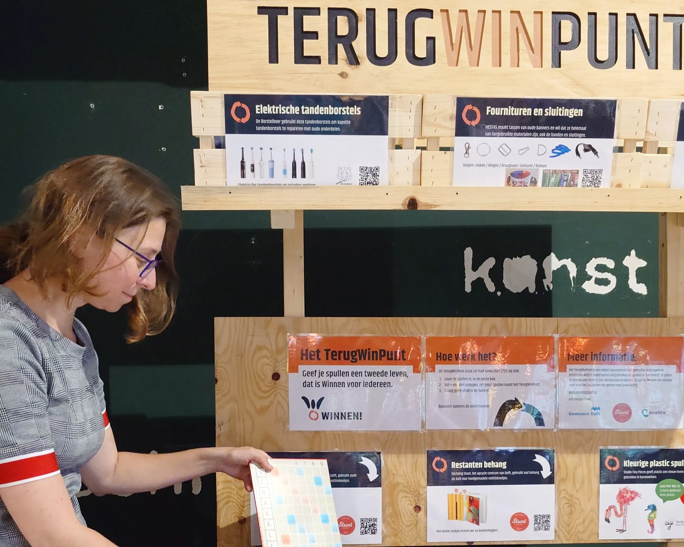Een vrouw met kort bruin haar en een bril staat voor een houten informatiebord. Op het bord staat in grote letters 'TERUGWINPUNT'. Daarnaast staat met krijt het woord 'kunst' op een groen vlak. Het bord bevat diverse kleine folders en flyers met informatie over recycling en duurzaamheid. De vrouw houdt een scrabble-bord vast en lijkt dit te bestuderen. Het geheel ziet eruit als een educatief punt gericht op duurzaamheid en afvalscheiding.
