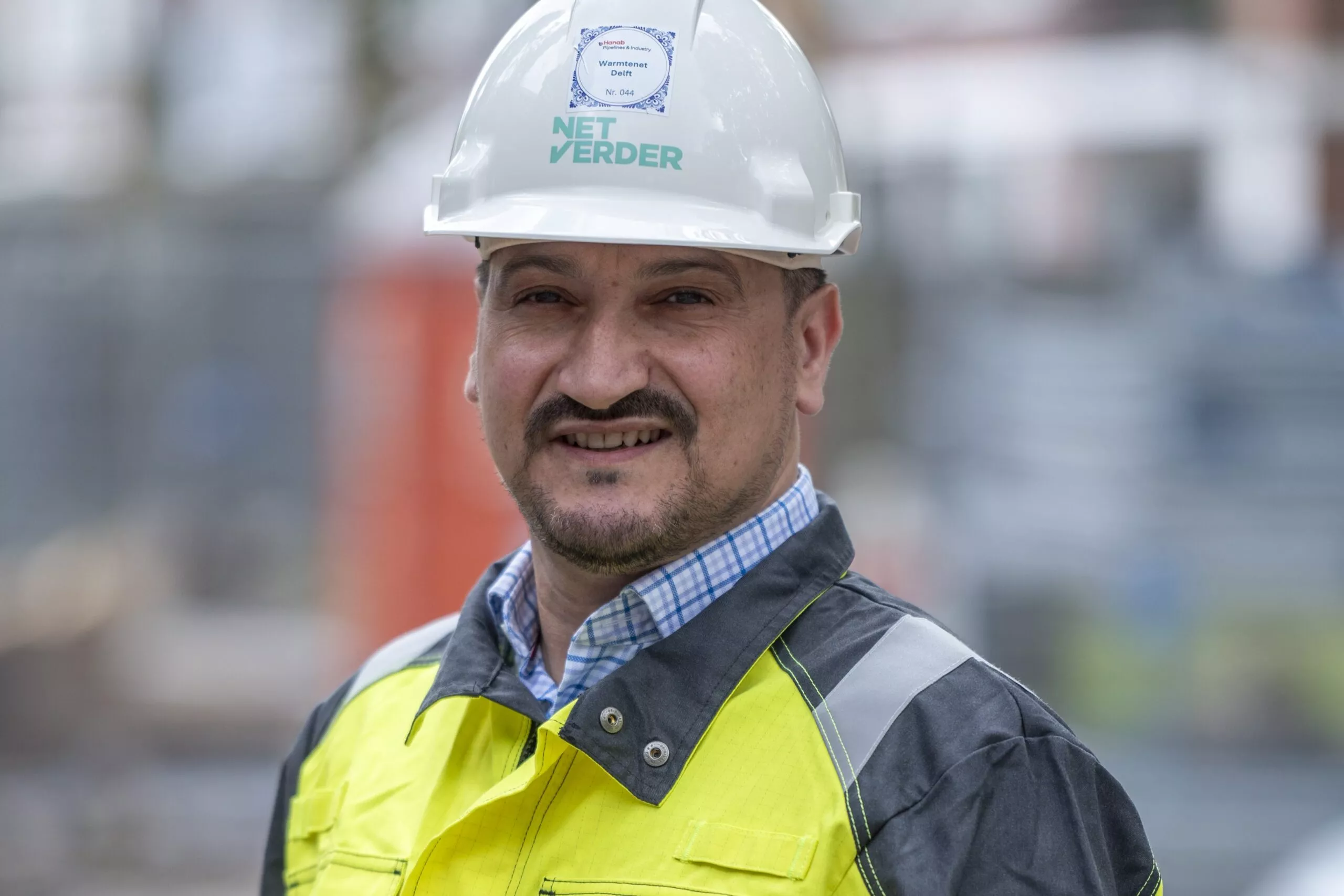 Een man met baard en snor draagt een witte bouwhelm en een opvallend geel-zwart veiligheidsjack met reflecterende strepen. Op zijn helm en jas staat tekst en een logo, er staat 