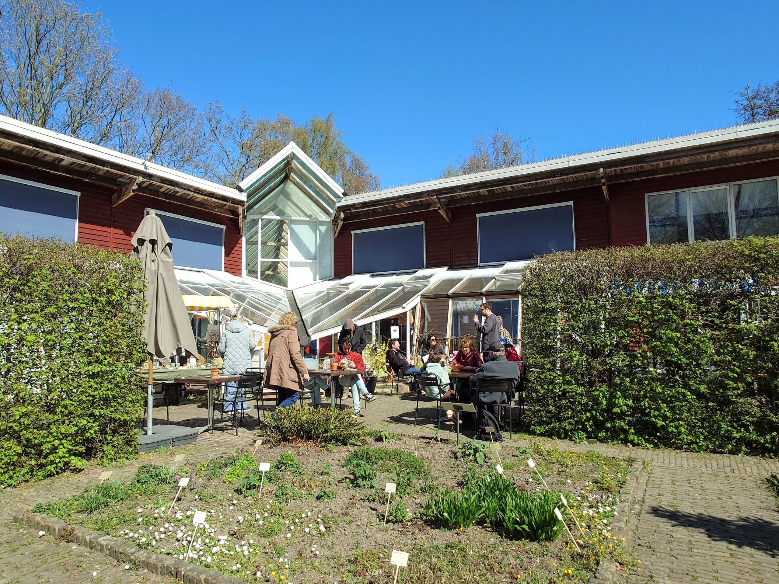 Een foto van een gebouw met een groot glazen terras/serre aan de voorkant, met daarvoor een terras waar meerdere mensen aan tafels zitten. Vooraan zie je een tuin/plantbed met kleine plantjes en bordjes; daarnaast een pad. In het midden staat het terras met stoelen en tafels en een grote, ingeklapte parasol. Achter het terras is een roodbruin gebouw met meerdere ramen en een glazen opbouw/serre. De lucht is helder blauw en er staan bomen op de achtergrond.