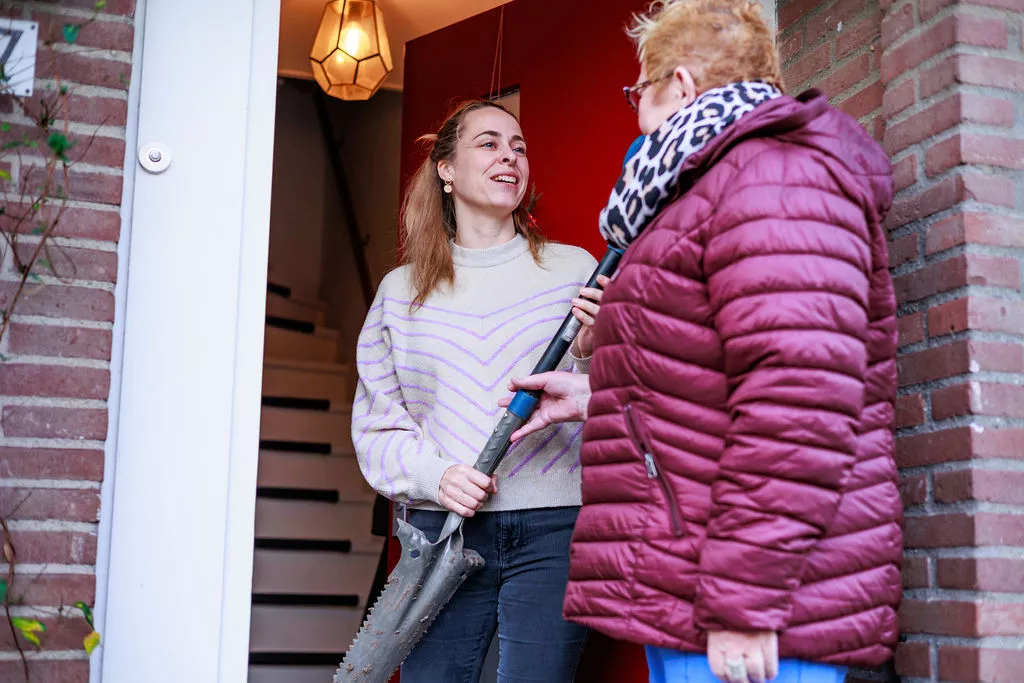 Een vrouw en een meisje bij een deuropening Beschrijving: - Voorgrond: een vrouw met kort blond haar en een paarse, gewatteerde jas met een rits en een sjaal met een zwart-wit motief. Ze staat met haar rug licht naar de camera en houdt met beide handen een lang metalen voorwerp vast. - Tweede persoon: een jongere vrouw met lichtbruin haar los, zij draagt een lichtgekleurde trui met dunne strepen en lacht terwijl ze het andere uiteinde van het metalen voorwerp vasthoudt. Ze staat binnen in de deuropening. - Achtergrond: binnenhuis met donkerrode muur en een warme hanglamp rechtsboven. Buitenzijde van de deuropening is baksteen. - Het metalen voorwerp lijkt op een lange steel met een zaag- of grijperdeel aan het uiteinde.