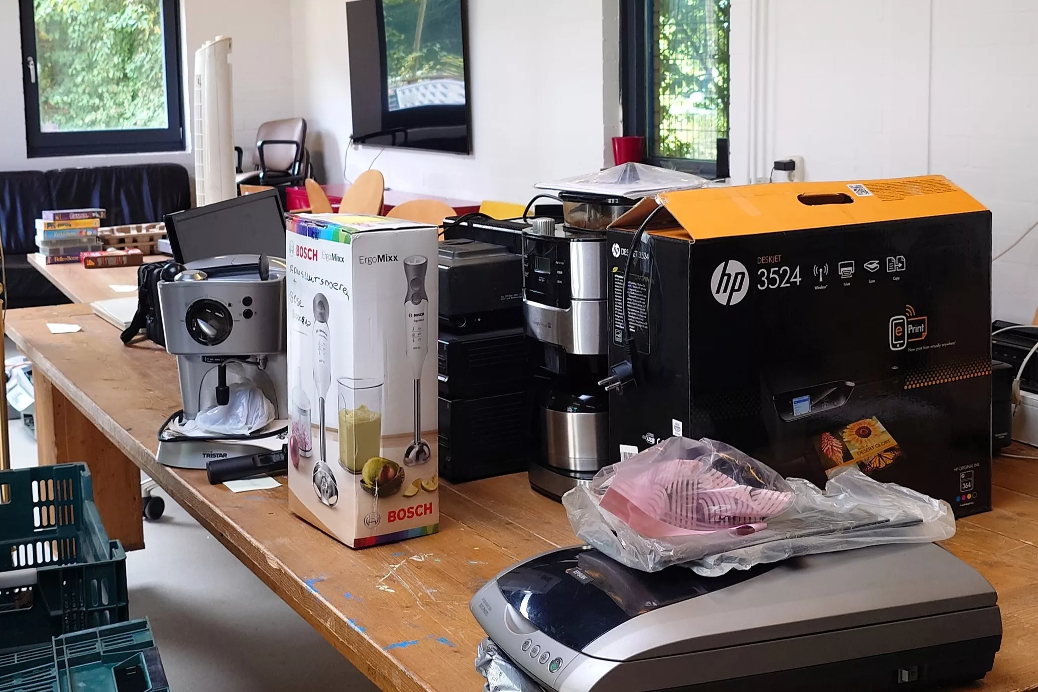 Ingeleverde keuken- en kantoorapparaten, zoals een espressomachine, printer, scanner en staafmixer, verzameld op een grote houten tafel voor recycling.