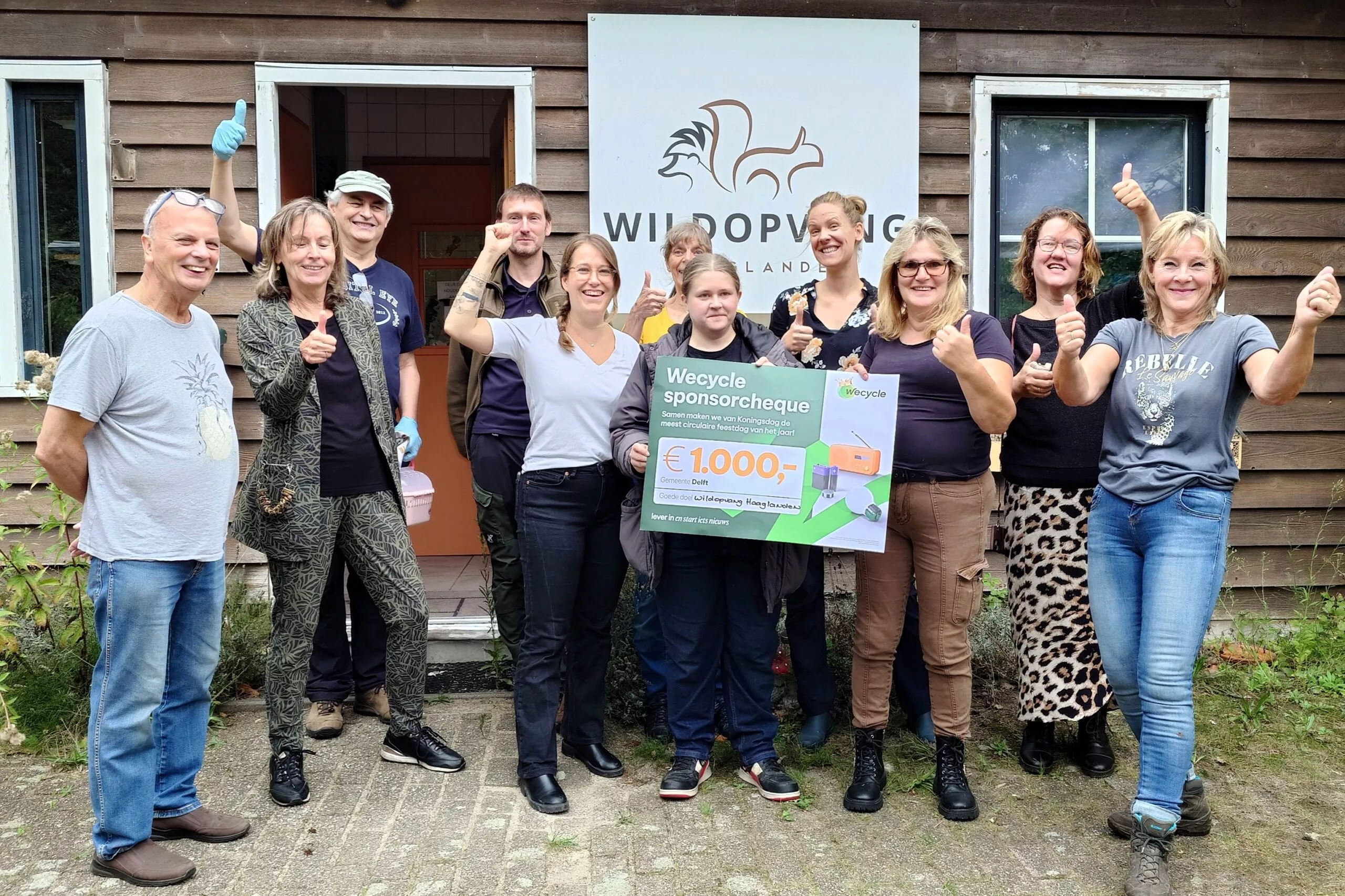Elf mensen staan vrolijk lachend met de duim omhoog voor het gebouw van Wildopvang Haaglanden, terwijl twee van hen in het midden een groene Wecycle sponsorcheque van € 1.000,- vasthouden.