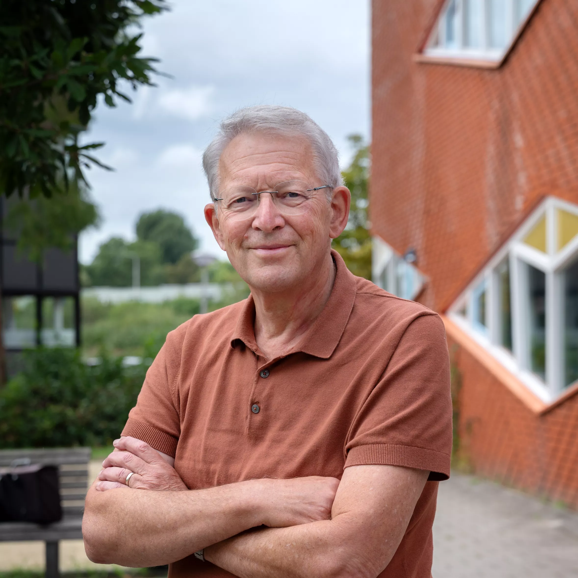 Glimlachende man met grijs haar, bril en roestbruine polo staat met gekruiste armen buiten voor een bakstenen gebouw met groen op de achtergrond.