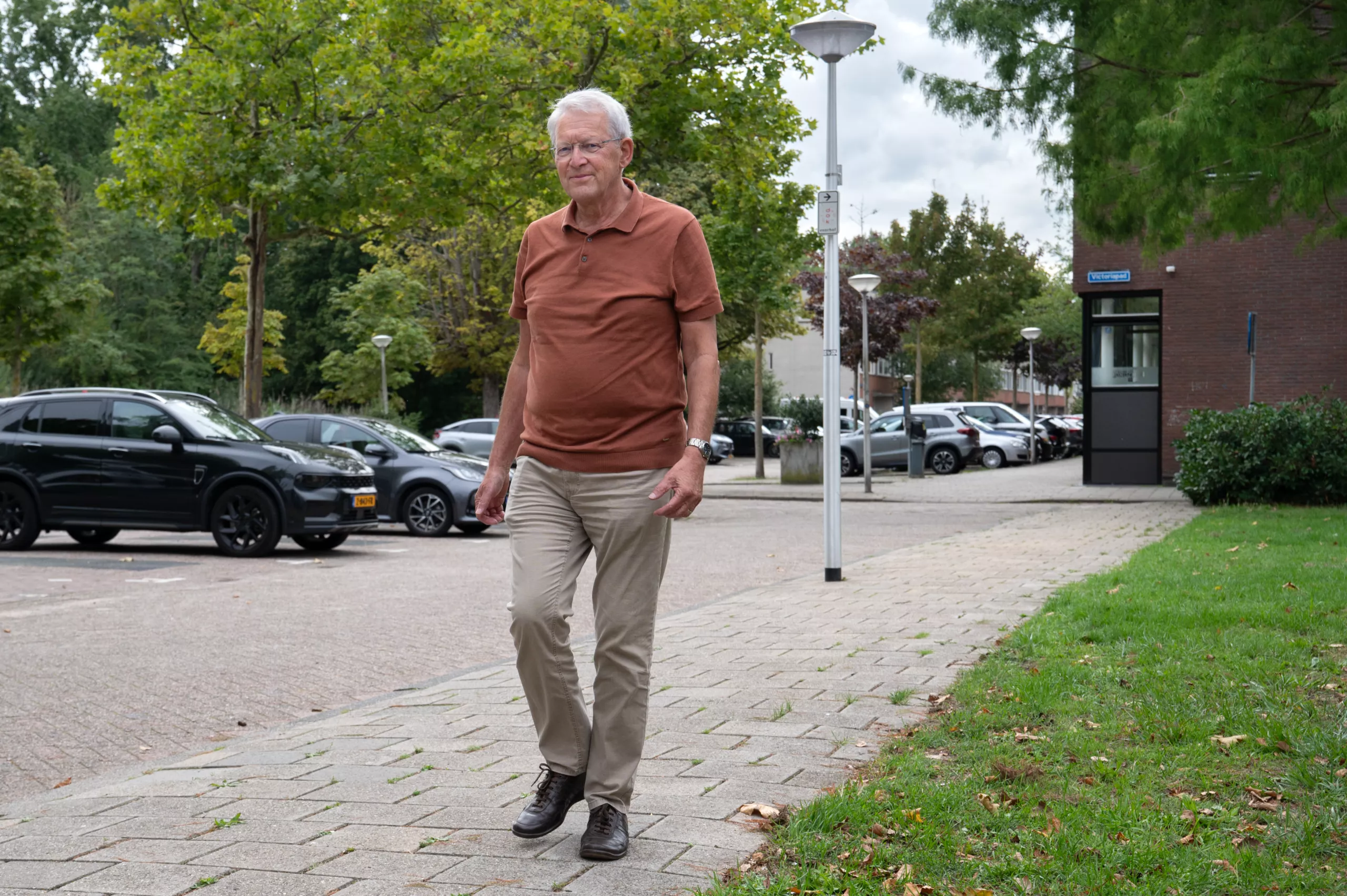 Oudere man met wit haar in roestbruine polo en beige broek wandelt over de stoep in een groene woonwijk.