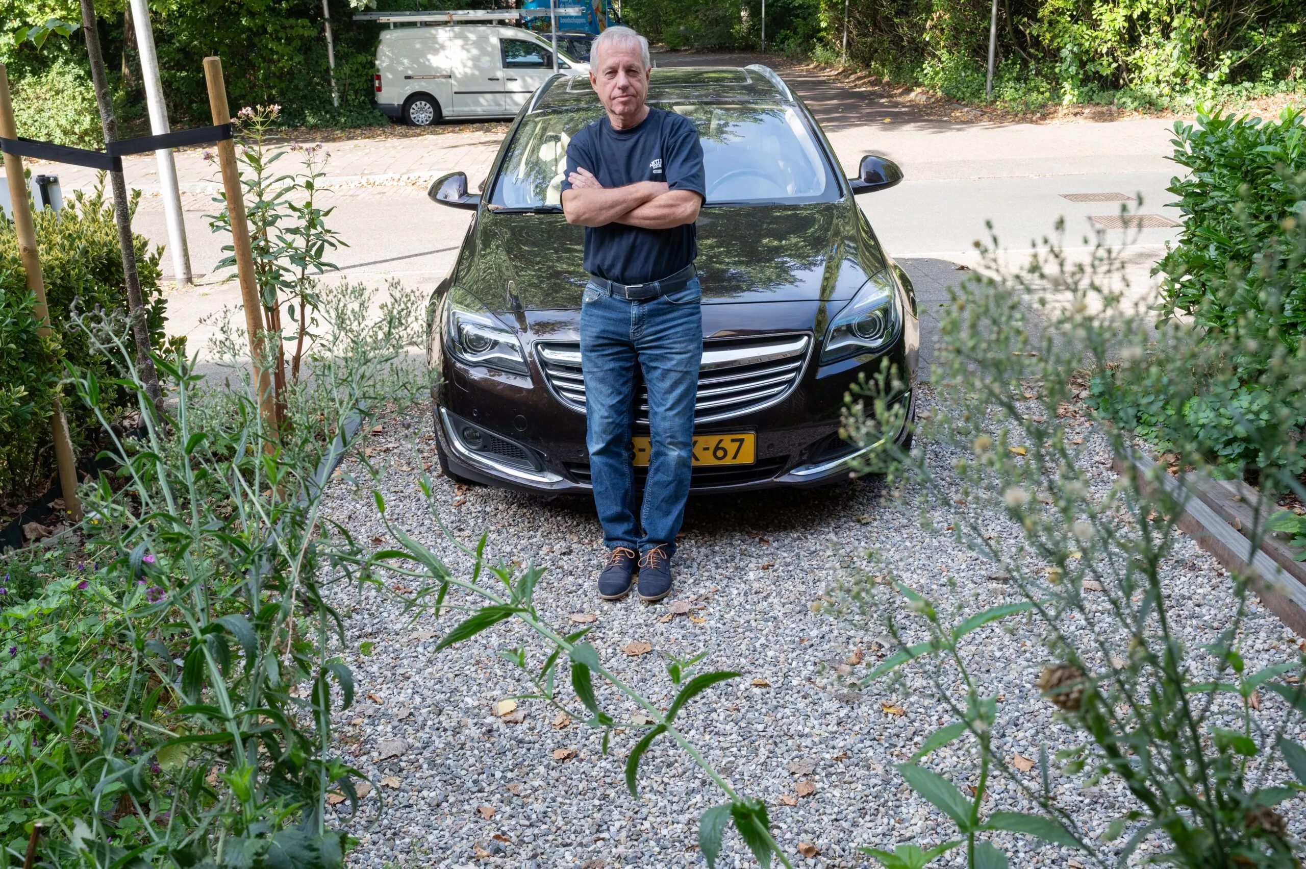 Een oudere man met grijs haar staat met gekruiste armen voor een glanzende zwarte sedan op een oprit van grind. Hij draagt een donker T-shirt en een spijkerbroek. De oprit is omgeven door groene planten en struiken, en op de achtergrond zijn bomen en een geparkeerde witte bestelbus te zien.