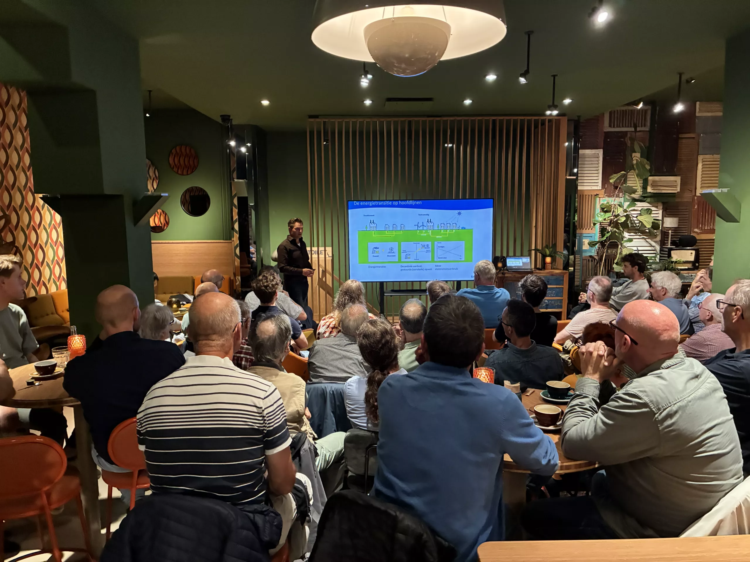 Een groep van twintig tot dertig volwassenen zit in een gezellig, klein zaaltje met groene muren en kijkt toe hoe een man een presentatie geeft bij een verlicht scherm. De bezoekers zitten met hun rug naar de camera aan tafels met kopjes en glazen. De sfeer is informeel en aandachtig.